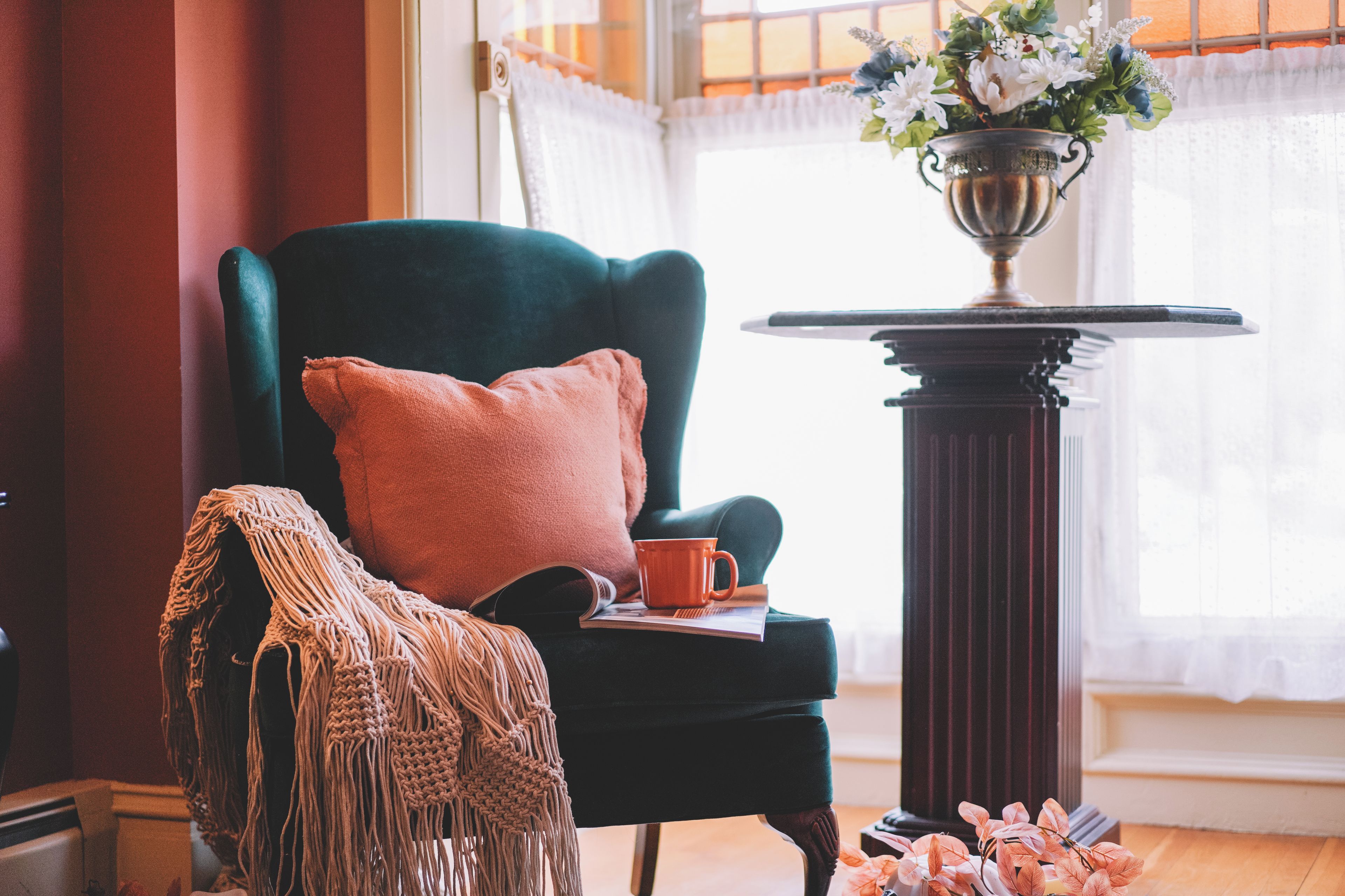 A room with a dark green armchair, a small table with a metal base, a cushion, a macrame throw, a book, and a mug.