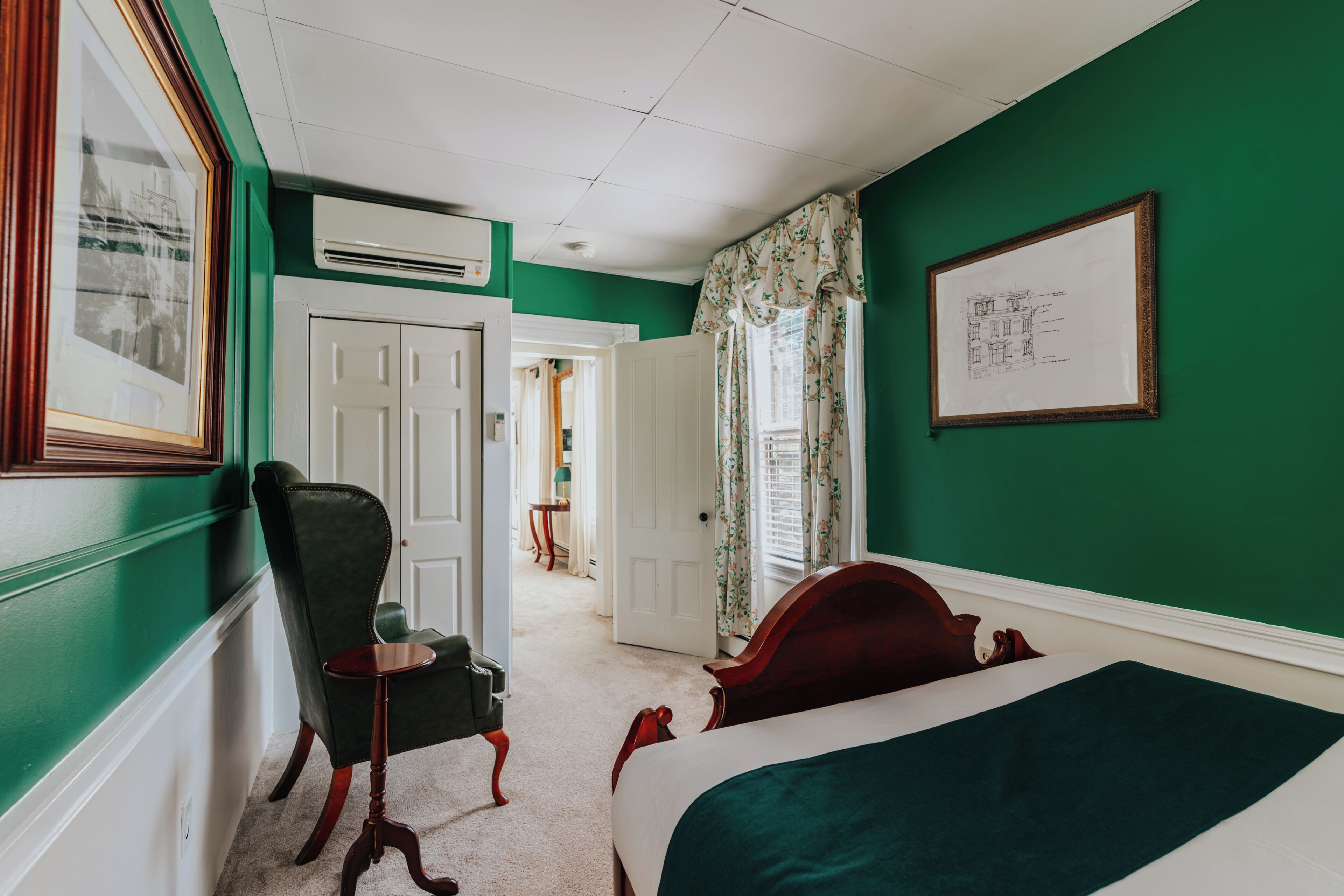 A hotel room with a bed, a chair, a desk, and a framed architectural drawing on the wall.