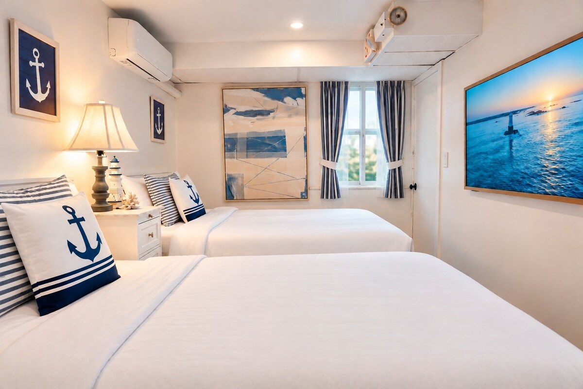 A hotel room with two beds, nautical-themed decor, a painting of a lighthouse, and a view of the sea.