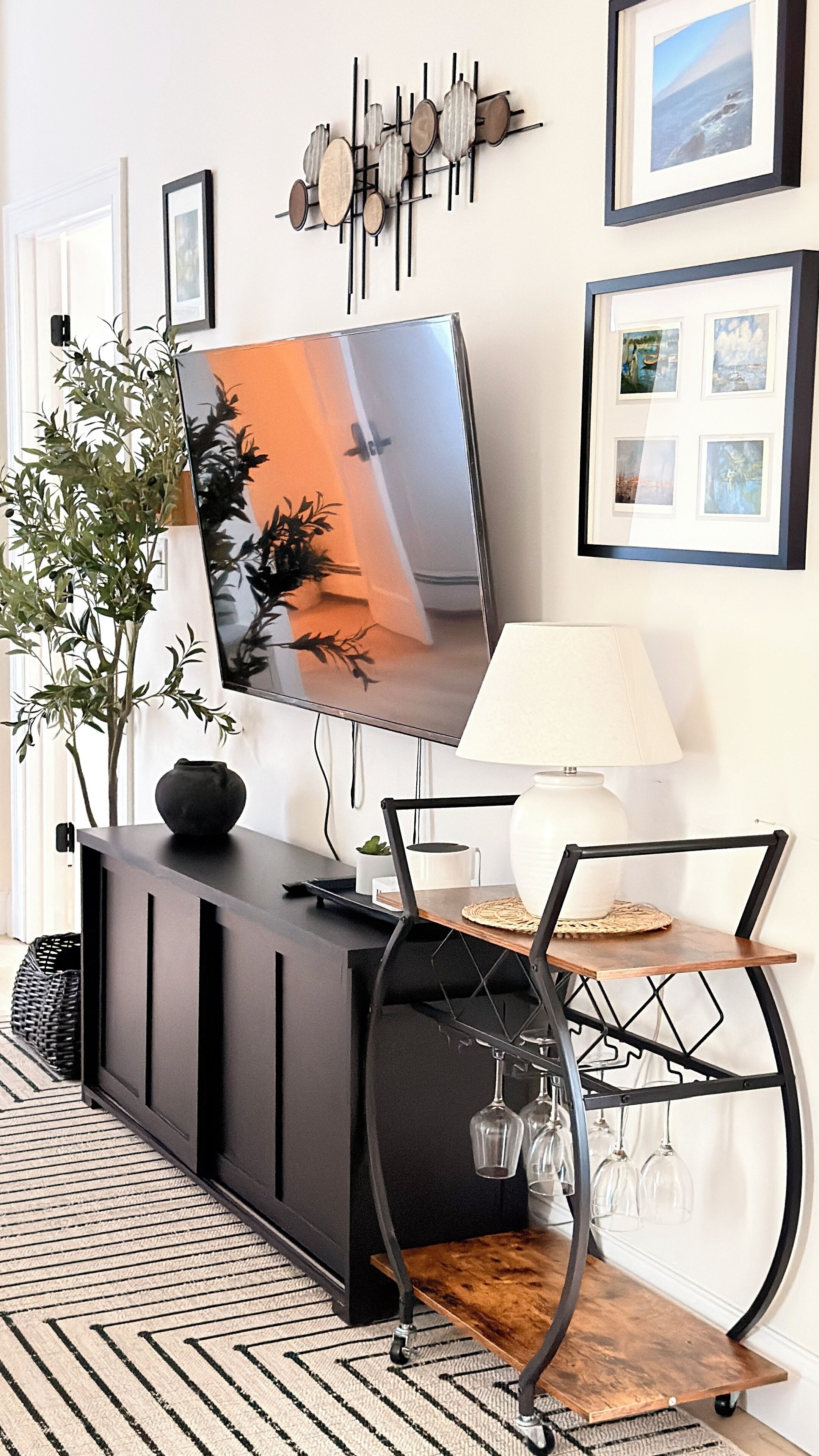 A modern living room with a flat-screen TV mounted on a black cabinet, a wooden coffee table with a glass top, a black metal chair, and a decorative wall piece above the TV.