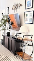 Una sala de estar moderna con un televisor de pantalla plana montado en un mueble negro, una mesa de centro de madera con tapa de vidrio, una silla de metal negro y una pieza decorativa en la pared sobre el televisor.
