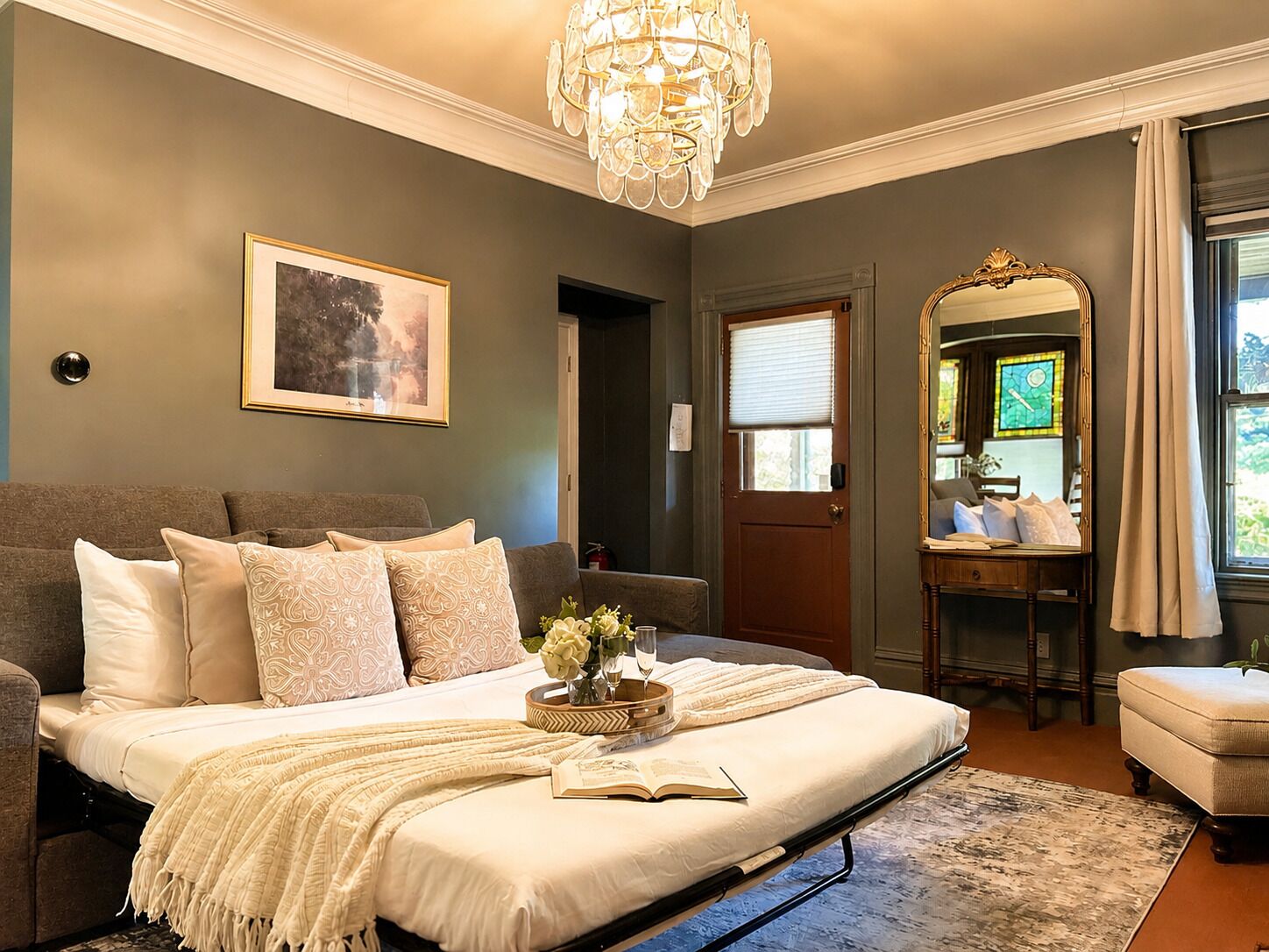 A bedroom with a bed, a chandelier, a framed picture, a mirror, and a door.
