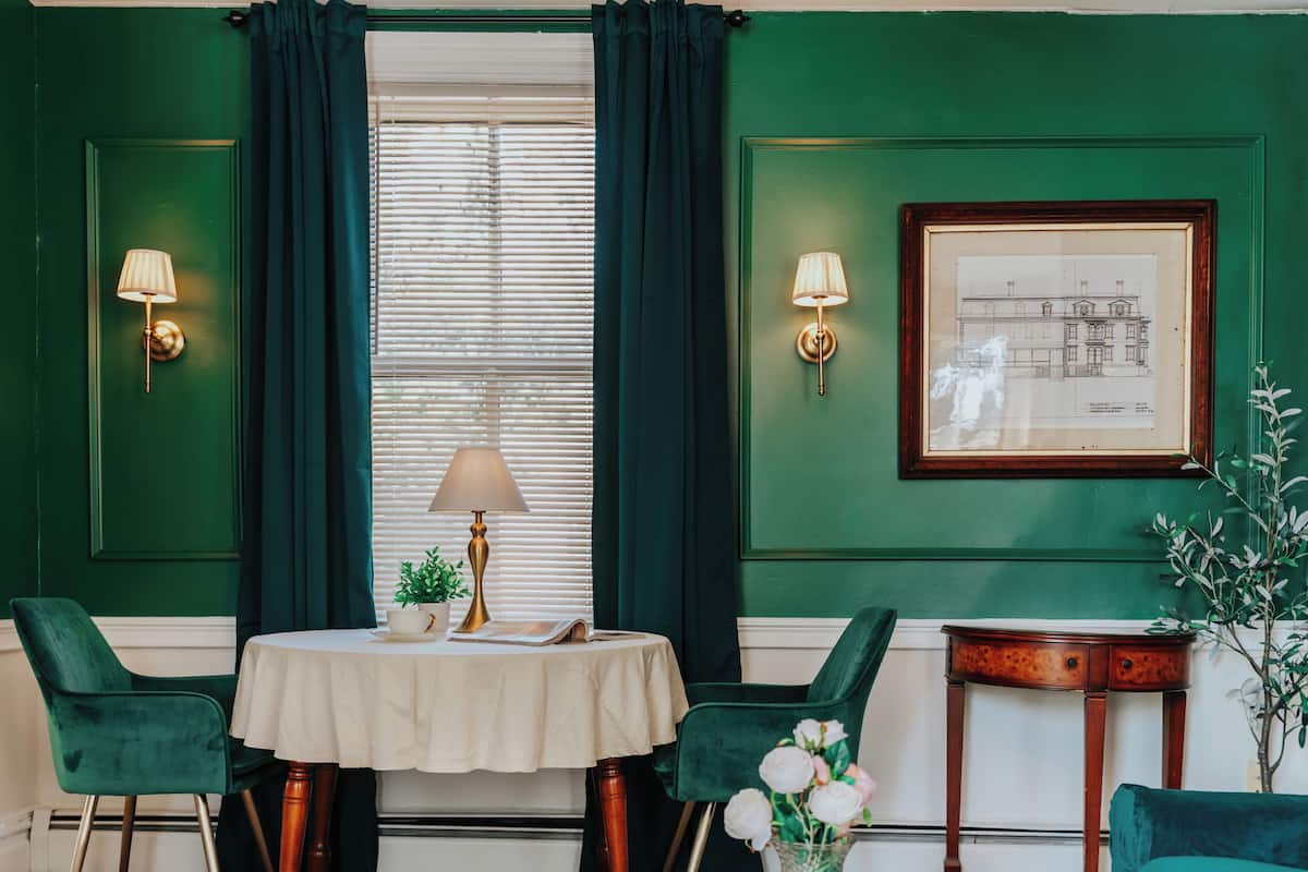 Habitación con paredes verdes, un dibujo arquitectónico enmarcado, dos lámparas fijadas a la pared, una mesa redonda con mantel blanco, dos sillas verdes y una pequeña mesita de madera con una planta.
