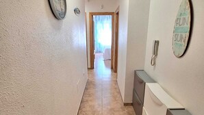 Un pasillo con piso de baldosas, una puerta de madera al final y un teléfono fijo en la pared.