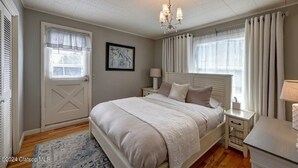 A bedroom with a large bed, a chandelier, a nightstand, a lamp, and a window with curtains. - 3 Bedroom Beach Oasis - HOT TUB - Pet Friendly (Seaside)