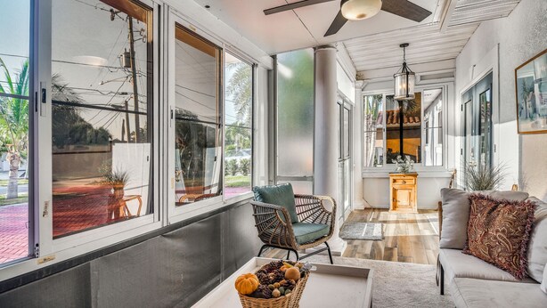 Une pièce spacieuse avec de grandes fenêtres, un ventilateur de plafond, un fauteuil en osier, un canapé et une petite table sur laquelle se trouve un panier.
