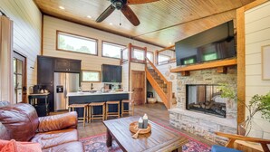 A spacious living area with a fireplace, leather sofa, and a kitchen in the background. - Tree-Lined Broken Bow Studio Cabin w/ Hot Tub! (Broken Bow)