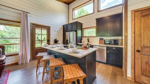 A kitchen with wooden cabinets, a large island with stools, and a view of the outdoors. - Tree-Lined Broken Bow Studio Cabin w/ Hot Tub! (Broken Bow)