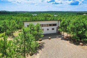 Une cabane de plain-pied avec une allée de gravier, entourée d’une forêt dense de pins, se dressant sur un fond de collines douces et sous un ciel dégagé.