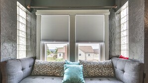 Un sofá gris con tres almohadones: uno verde azulado, dos estampados de leopardo y uno rojo, ubicado frente a una ventana con persianas cerradas.