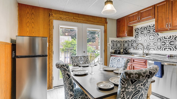 A kitchen with a stainless steel refrigerator, wooden cabinets, patterned backsplash, and a dining table set with plates and glasses. - 11 Mi to Niagara Falls: North Tonawanda Apartment (North Tonawanda)