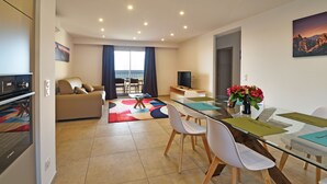 A modern living room with a sofa, TV, and dining area with chairs and a glass table. - Résidence Saint-jean : Appartement Agriate - RDC - Vue mer (Saint-Florent)