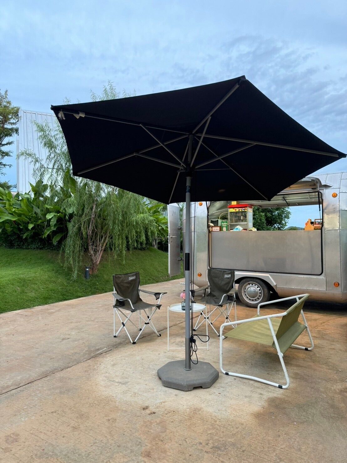 Un espace extérieur aménagé avec une table et des chaises sous un grand parasol, un barbecue portable et un camion-restaurant en arrière-plan.