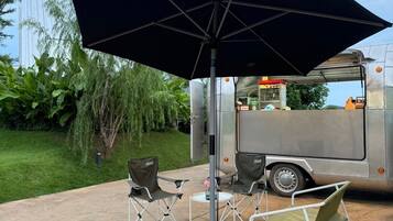 Un espace extérieur aménagé avec une table et des chaises sous un grand parasol, un barbecue portable et un camion-restaurant en arrière-plan.