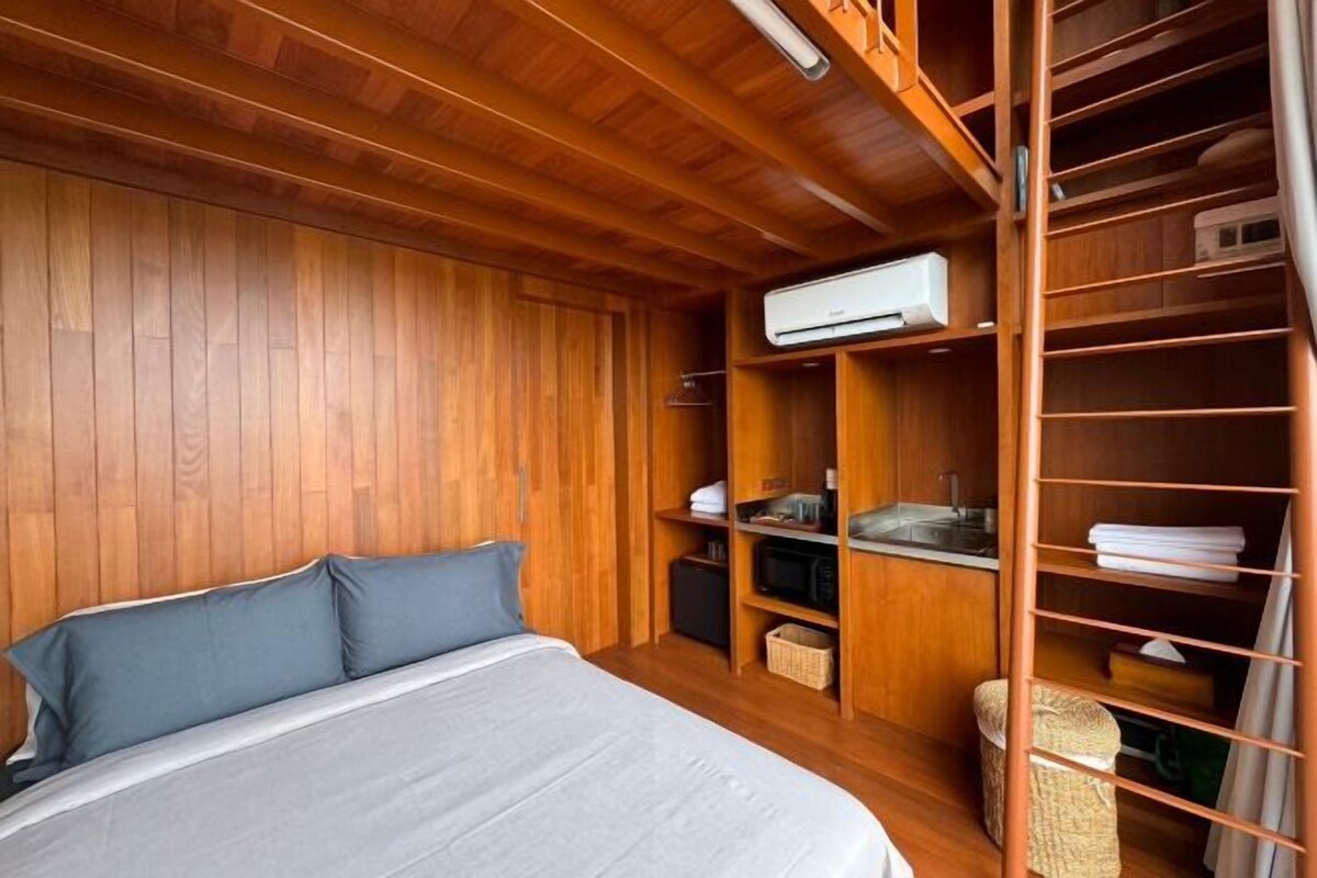 A bedroom with a wooden ceiling, a bed with grey bedding, a built-in wardrobe, a microwave, and a small sink.