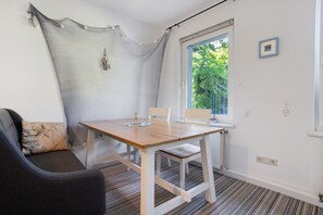 Une pièce avec une table en bois, des chaises blanches, un canapé gris, une fenêtre donnant sur un paysage verdoyant et un mur blanc orné d’un cadre.