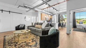 Ein modernes Wohnzimmer mit einer Couch, Sesseln und einem Couchtisch. Im Hintergrund ist eine Küche mit Schränken und einer Mikrowelle zu sehen.