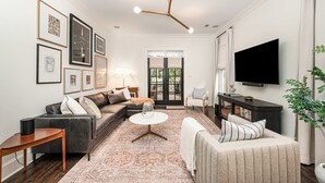 Una sala de estar moderna con un sofá de cuero oscuro, una mesa de centro redonda y un sillón color beige. Hay un televisor grande montado en la pared, una araña y obras de arte enmarcadas.