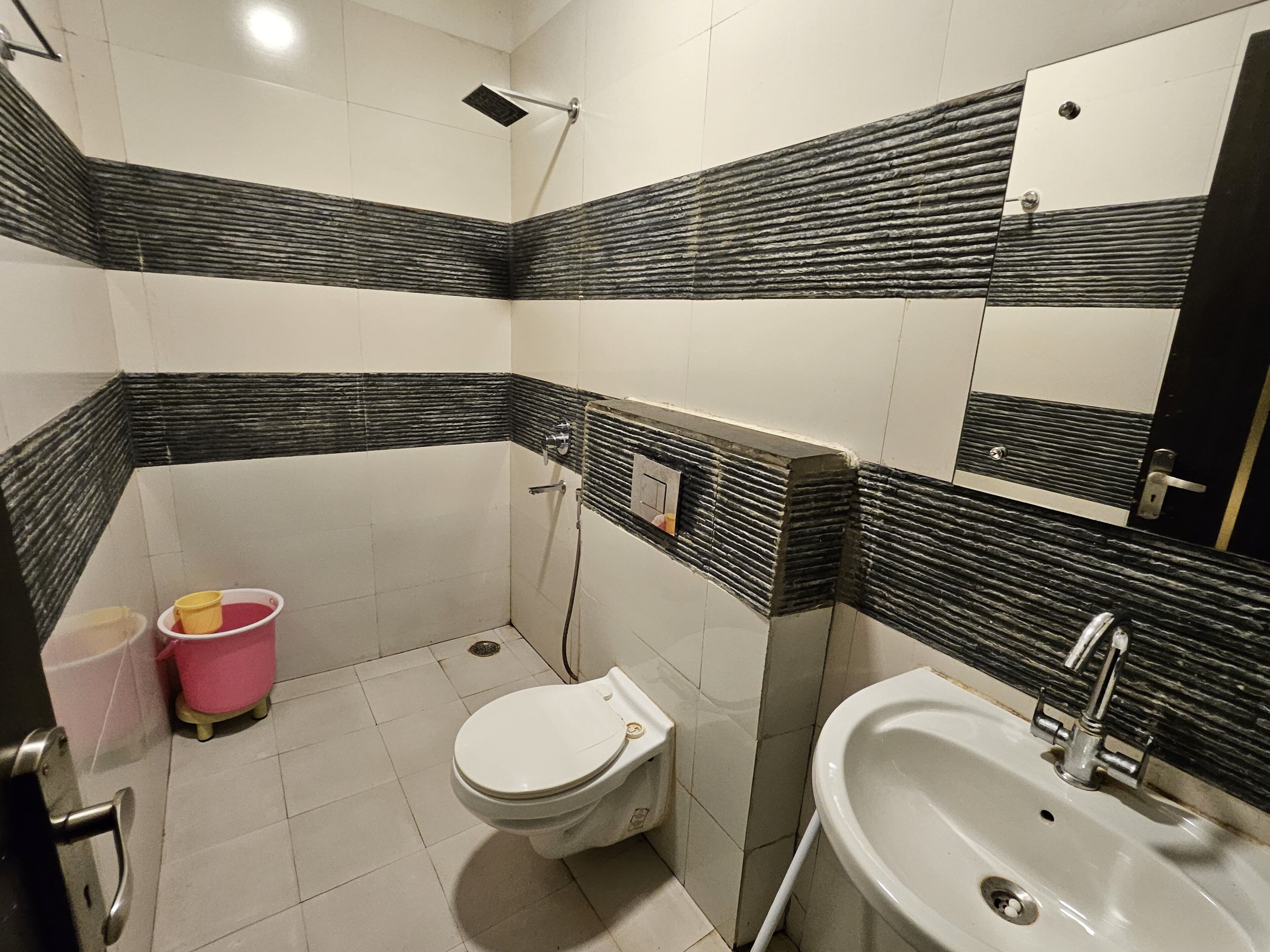 A bathroom with a toilet, sink, and shower. There are two buckets, one pink and one white, on the floor.