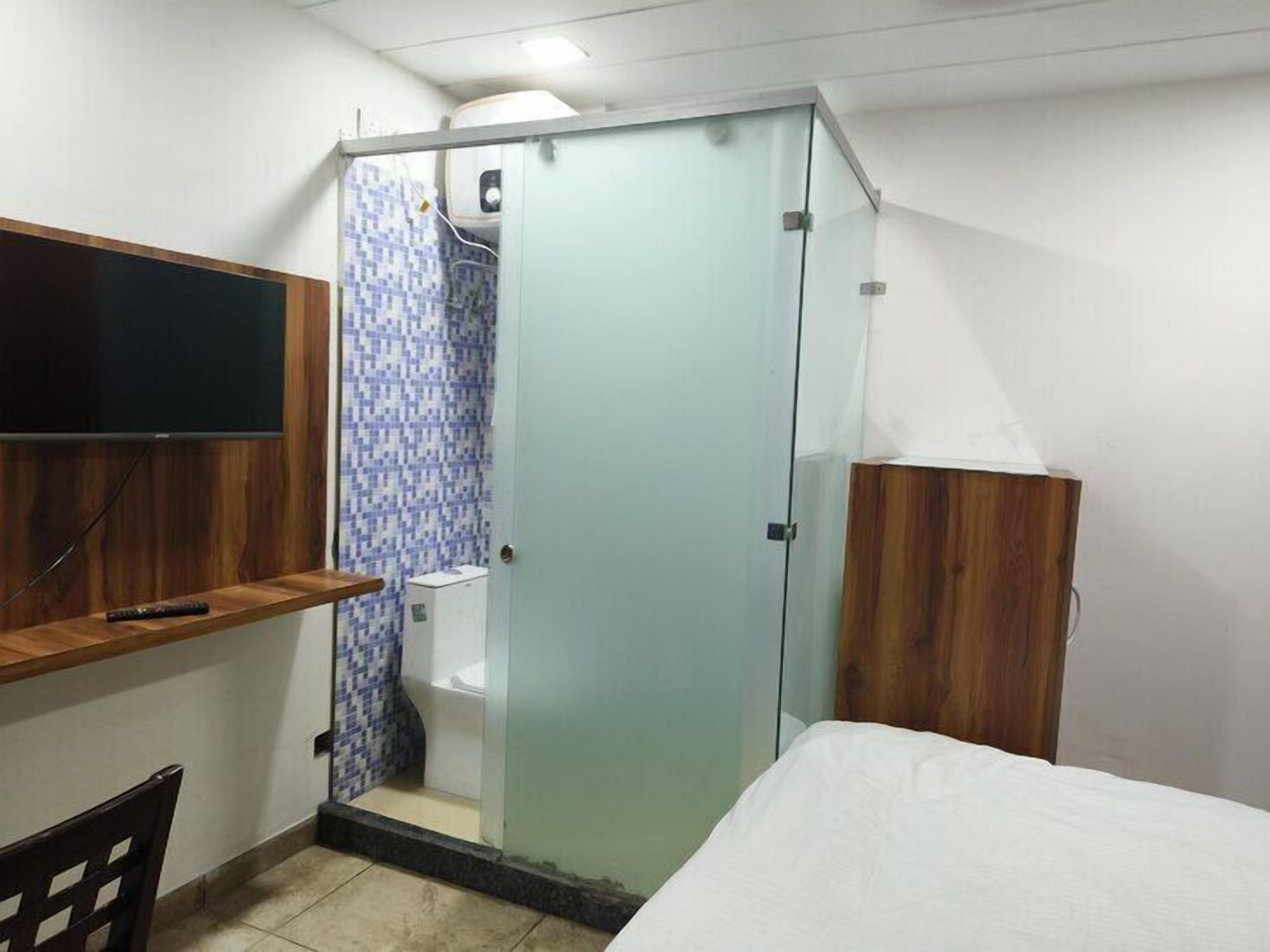 A small, compact room with a bed, a wooden desk, a television, and a shower area with a tiled wall.