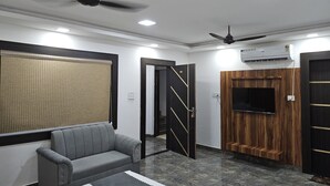 A modern living room with a grey sofa, a ceiling fan, and a flat-screen TV mounted on a wooden panel wall. - Jhansi Castle Inn (Jhansi)