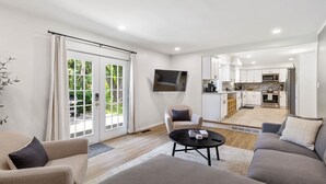 A modern living room with a sofa, armchair, and coffee table, a kitchen in the background, and a sliding glass door leading to a balcony. - Upscale 6BR Home w/ Office (Clinton Township)