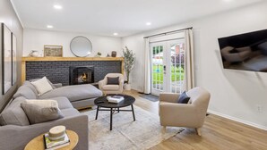 A modern living room with a fireplace, a sofa, a coffee table, and a television on the wall. - Upscale 6BR Home w/ Office (Clinton Township)