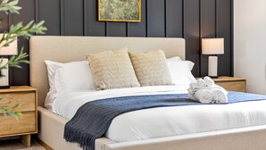 A neatly made bed with white linens and two beige pillows, a blue throw blanket, and a stack of white towels on a wooden nightstand. - Upscale 6BR Home w/ Office (Clinton Township)