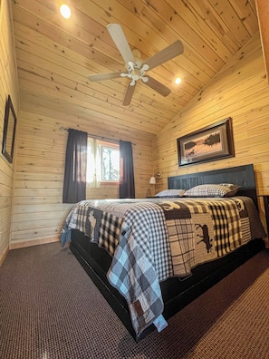 A wooden cabin bedroom with a bed, a ceiling fan, a window with curtains, and a framed picture on the wall.