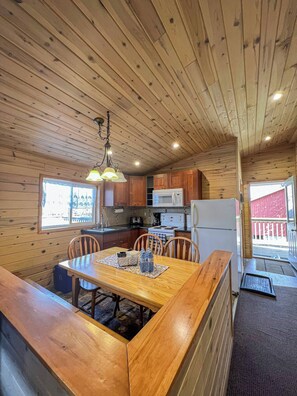 A wooden cabin interior with a dining table, chairs, kitchen area, and a window.