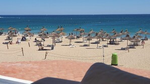 Een strand met rijen rieten parasols en ligbedden, een helderblauwe lucht en de oceaan op de achtergrond.