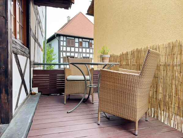 Eine Holzterrasse mit Korbmöbeln, einem Blumentopf und Blick auf ein traditionelles Gebäude.