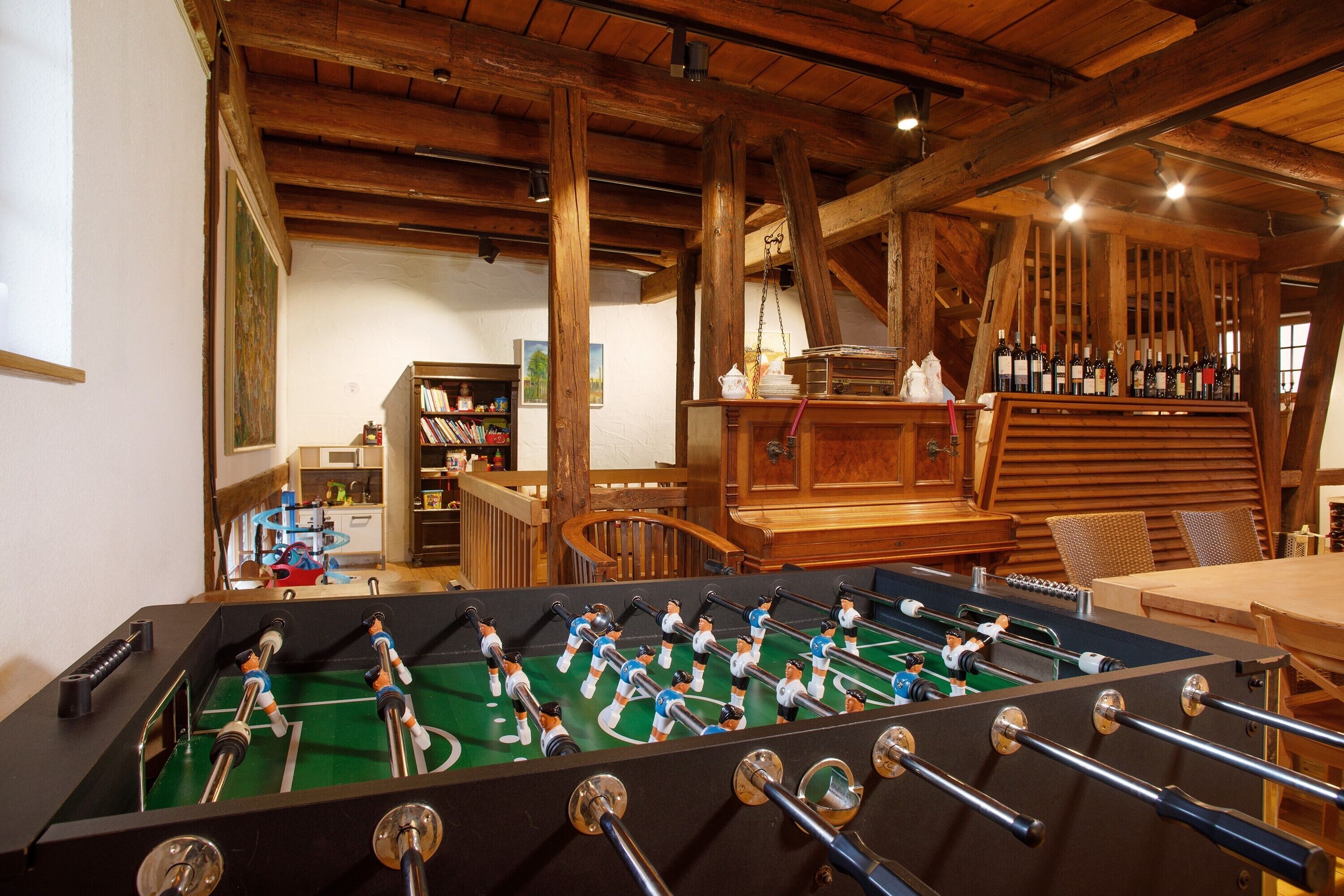 A room with a foosball table, wooden beams, a kitchen area, and a bookshelf.