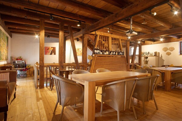 Een rustieke eethoek met houten balken, een lange tafel, rieten stoelen en een wijnrek.