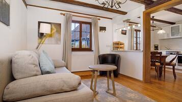 Ein gemütliches Wohnzimmer mit einer Couch, einem kleinen runden Tisch und einem Sessel. Ein Fenster mit Vorhängen, ein Bild an der Wand und ein durchgehender Blick in eine Küche.