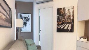 Un salon moderne avec un canapé gris, une petite table et un climatiseur fixé au mur. Deux œuvres d’art encadrées ornent le mur : l’une représente un paysage urbain et l’autre, un gros plan d’une main.