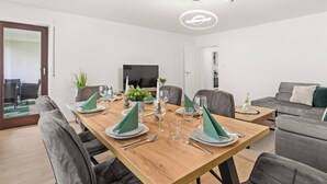 Ein moderner Essbereich mit einem Holztisch, gedeckt für vier Personen, umgeben von grauen Polsterstühlen. An der gegenüberliegenden Wand ist ein Flachbildfernseher angebracht.