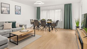 Ein modernes Wohnzimmer mit einem grauen Sofa, einem Holztisch, einer Essecke mit Tisch und Stühlen sowie einem großen Fenster mit grünen Vorhängen.