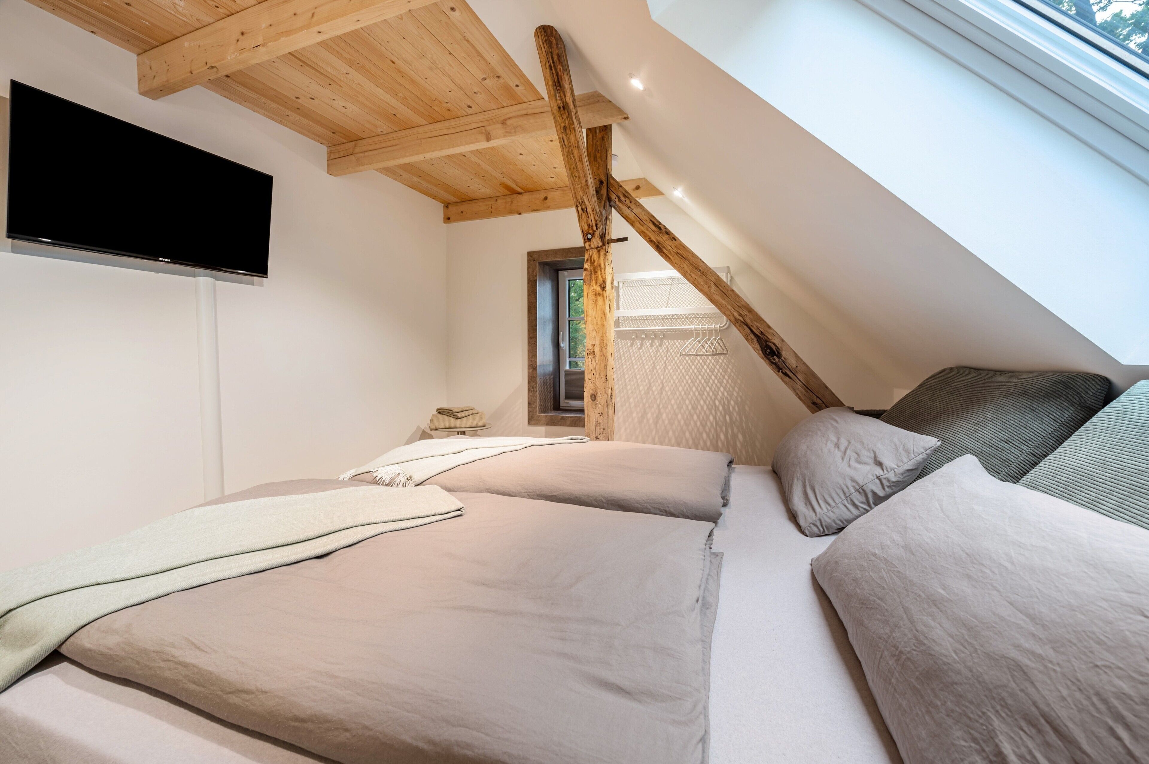 Et soveværelse med to senge, et fjernsyn ophængt på væggen og et skråt loft med træbjælker.