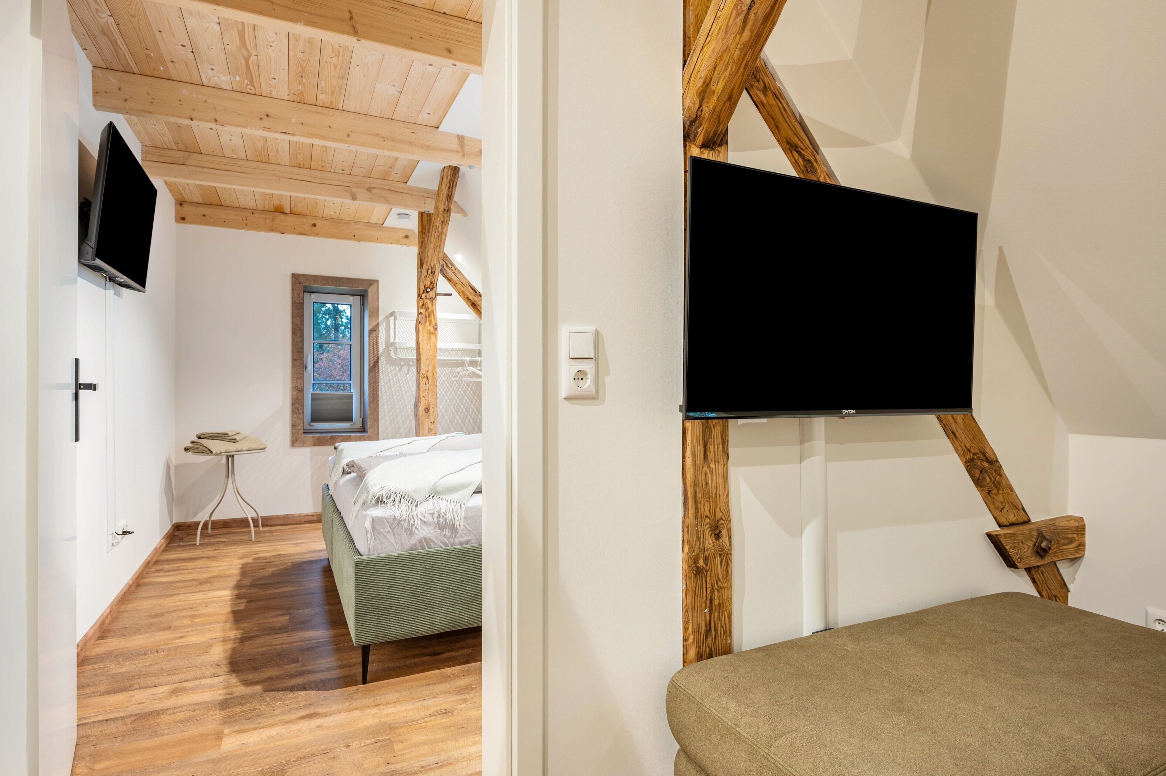 Een kamer met houten plafond, een bed, een klein tafeltje, een aan de muur bevestigde televisie en een raam met uitzicht.