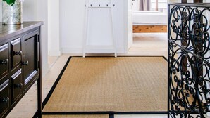 Ein Flur mit einer weißen Tür, einem schwarzen Metallgeländer, einer gewebten Matte und einem Bett mit Holz Kopfteil im Hintergrund.