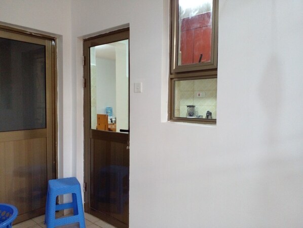 Una habitación con una puerta de madera, un taburete de plástico azul y una ventana que da a un objeto rojo en el exterior.