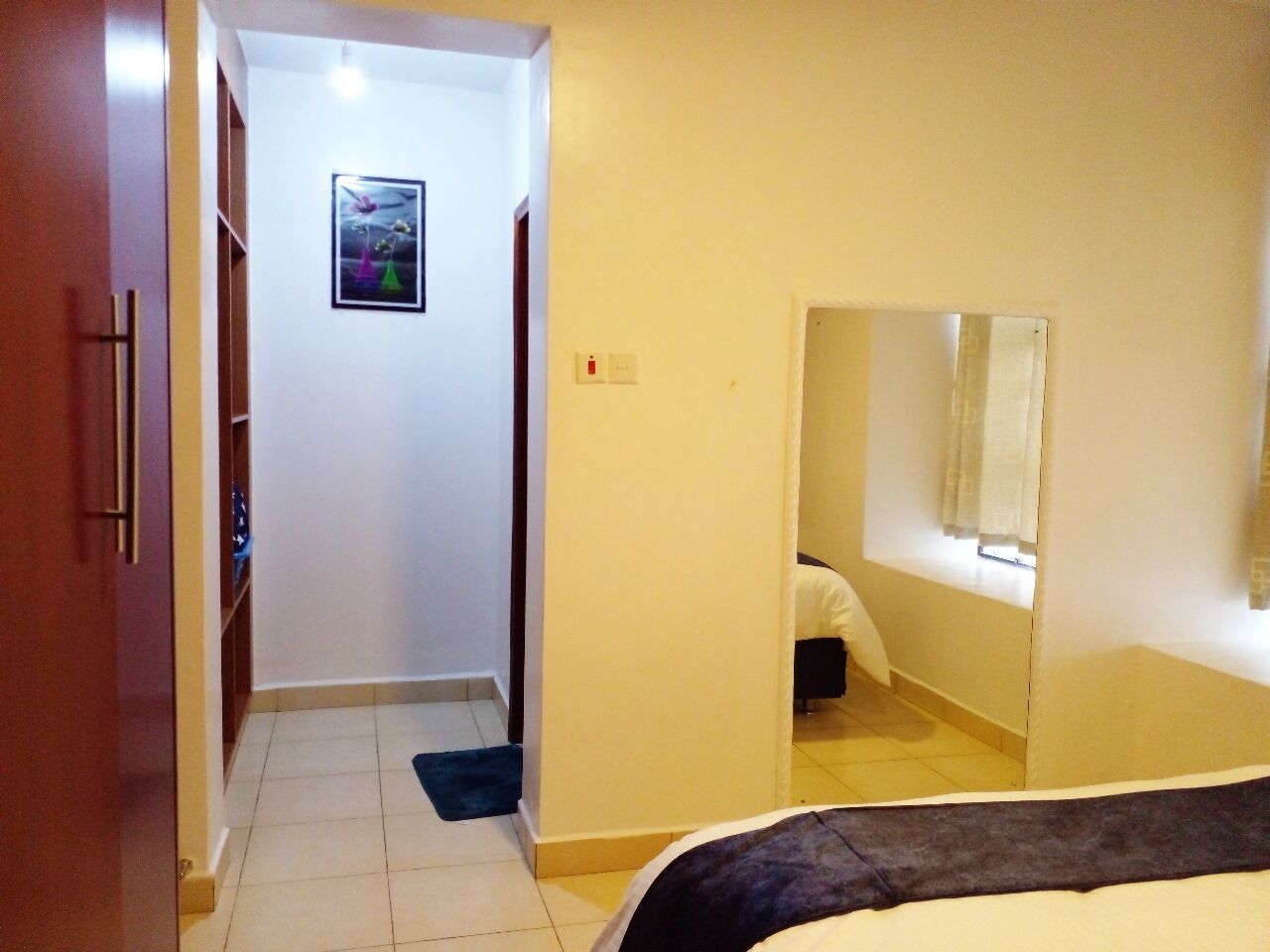 A hotel room with a bed, a wardrobe, a bathroom with a mirror, and a hallway leading to another room.