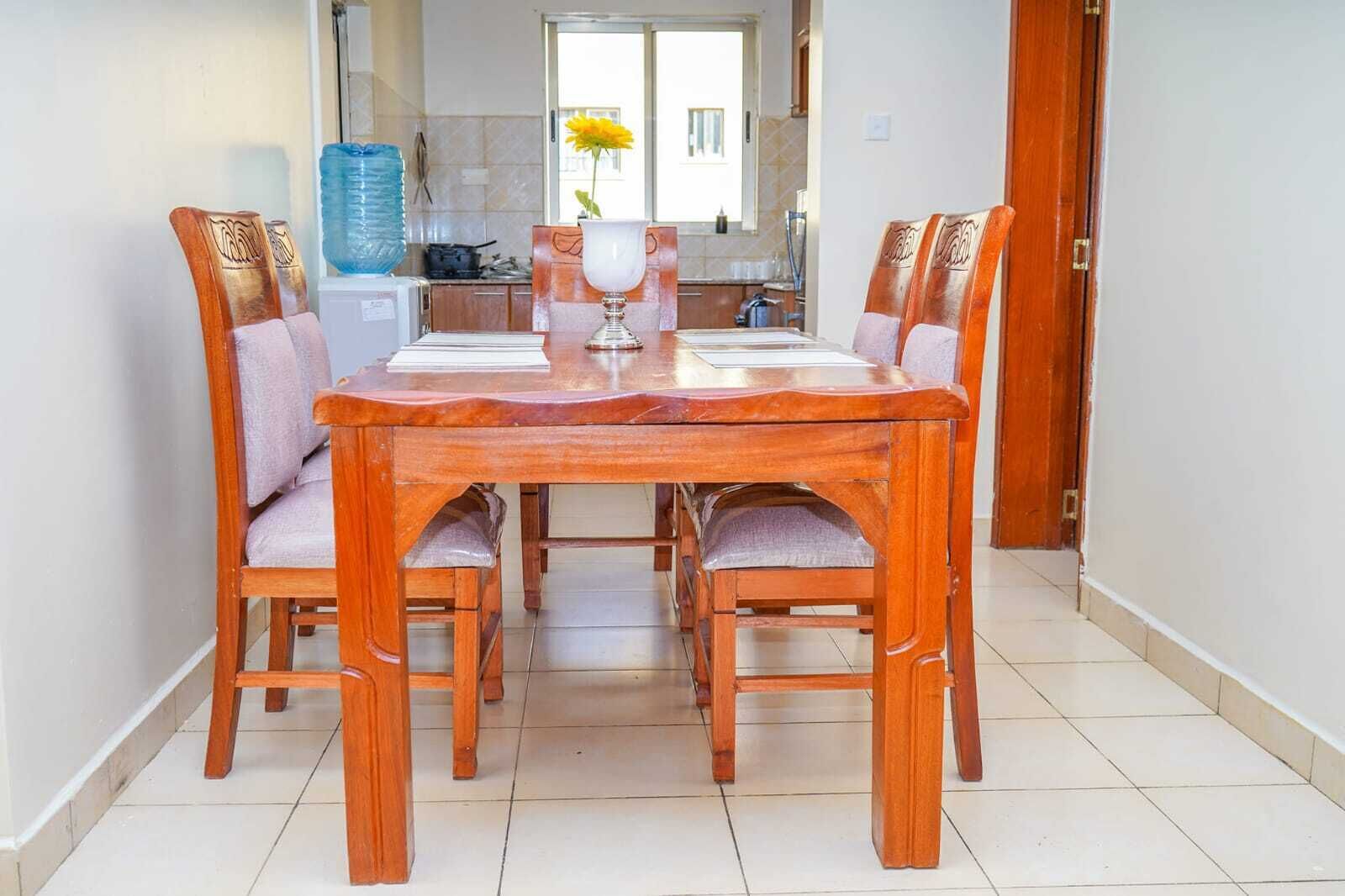 A dining area with a wooden table and chairs, a vase with a yellow flower, and a kitchen in the background.