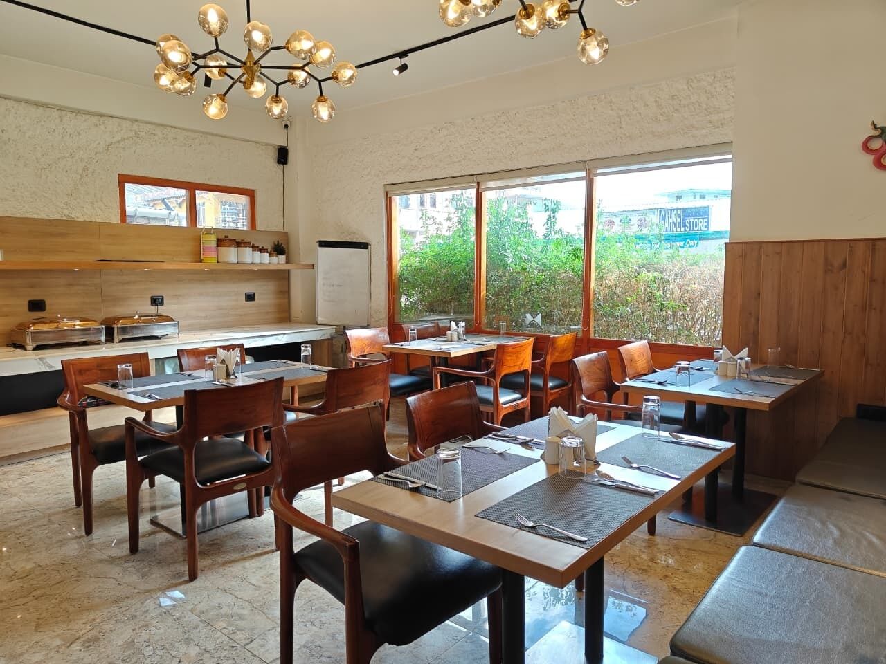 Un restaurante con varias mesas y sillas, una zona de cocina y grandes ventanales con vistas a zonas verdes.