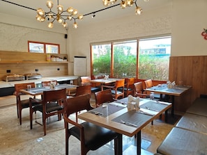 A restaurant with multiple tables and chairs, a kitchen area, and large windows with a view of greenery. - Hotel Dralha  (Thimphu)