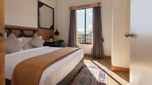 A bedroom with a large bed, a desk, a chair, a window with curtains, and a view of buildings. - Hotel Dralha  (Thimphu)