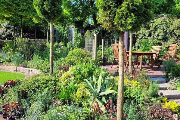 A garden with a wooden table and chairs, surrounded by various plants and a stone pathway.