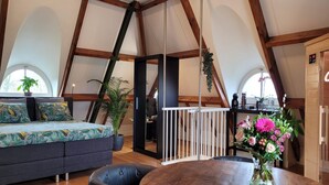 A loft-style room with a high ceiling, exposed wooden beams, a sofa with patterned cushions, a dining table with a vase of flowers, and a view through a window.
