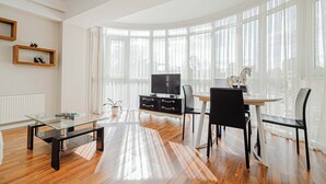 Ein modernes Wohnzimmer mit Glastisch, schwarz-weißen Möbeln, einem Fernseher und großen Fenstern mit durchsichtigen Vorhängen.
